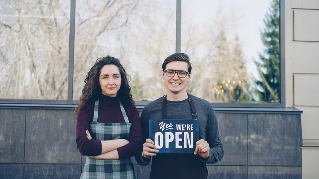 Two small business owners in aprons holding a Yes We're Open sign outside their shop